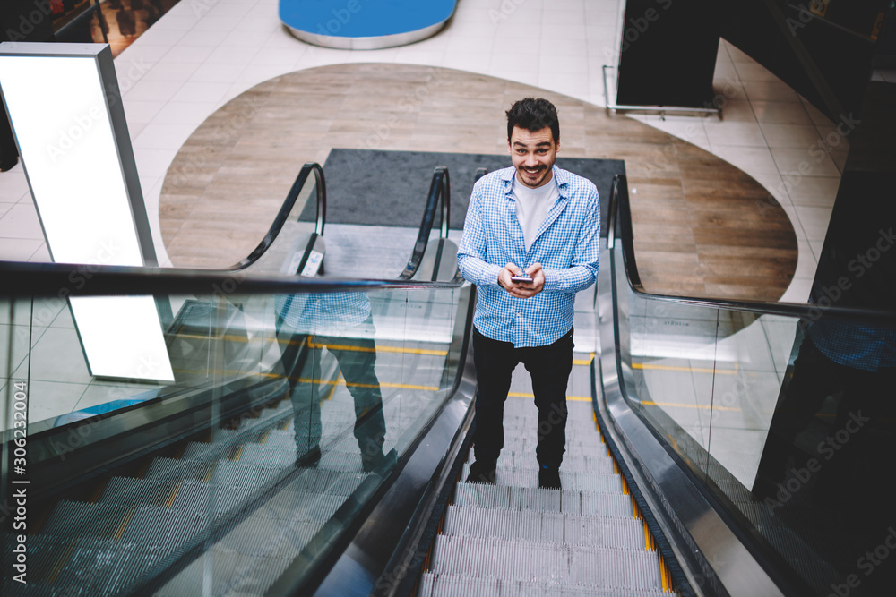 Poster portrait of cheerful hipster guy standing on escalator with smartphone gadget in hands and smiling a