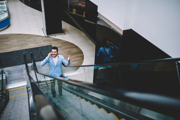 Happy excited male enjoying communication via cellular phone while spending time in shopping center, positive cheerful hipster guy on electric stairs calling to friend via application on cellphone