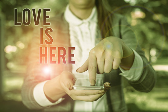 Word writing text Love Is Here. Business photo showcasing Roanalysistic feeling Lovely emotion Positive Expression Care Joy Woman in grey suites holds mobile phone