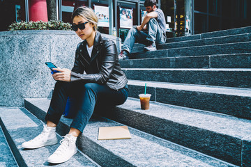 Serious chinese woman in sunglasses checking mail reading messages on mobile sitting on stairs...