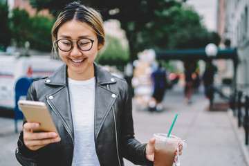 Smiling Asian woman using mobile application for strolling in downtown holding coffee to go, pretty Chinese hipster girl in trendy wear receive message while walking on street enjoying work break