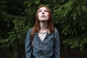 A redhead girl with blue eyes in a denim shirt stands straight and looks up holding her head against the background of fir trees