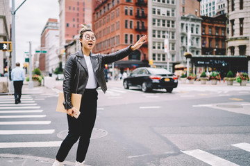 Young charming emotional asian woman in glasses for better vision gesturing while standing near to road for stopping taxi.Pretty hipster girl shouting while trying catch public transport outdoors