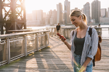 Fototapeta na wymiar Cheerful millennial female wanderer with touristic backpack searching online article about American vacation connecting to roaming internet for browsing websites on cellphone, international messaging