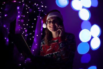 girl student in Christmas clothes sitting at night with a laptop and talking on the phone, she communicates online at Christmas