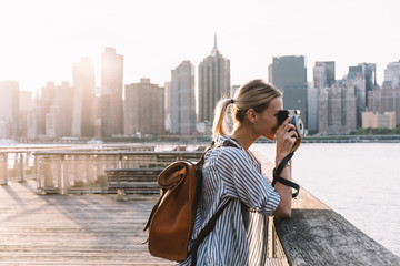 Side view of skilled hipster girl with instant camera clicking picture of city landscape standing at urban setting with Manhattan view, positive female tourist with backpack enjoying getaway