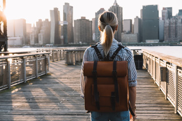 Back view of stylish female tourist with traveling backpack standing on American urban setting and examanise landmark of Manhattan district, millennial woman exploring United States during journey