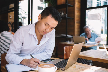Adult male working remotely writing notes while using laptop in cafe