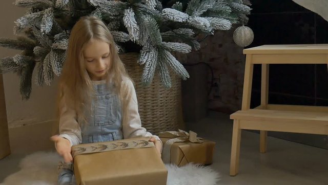 Cute blond girl has recieved a gift box wrapped in craft paper sitting near christmas tree, happy childhood and xmas celebration concepts