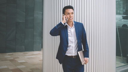 Confident young modern Asian businessman with laptop standing and speaking over smartphone