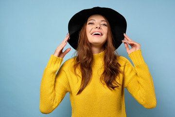 A brunette in a yellow sweater stands against a blue background, laughs with her head thrown back and holds the hat with her hands