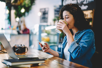 Caucasian brunette woman dressed in casual wear enjoying caffeine beverage during messaging with followers from own web blog connected to 4g wireless on smartphone device for browsing internet