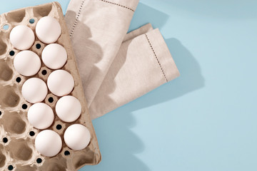 Chicken eggs in a white cardboard container lie on blue wooden kitchen table. One egg is broken. A kitchen towel lies nearby