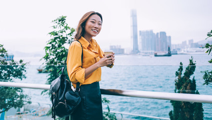 Fototapeta na wymiar Half length portrait of cheerful Asian tourist with coffee to go in hand standing near river and metropolis landscape on background smiling and enjoying free time for coffee to go in Hong Kong