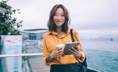 Half length portrait of millennial Japanese blogger with digital gadegt and coffee to go in hands smiling at camera during time for communication with virtual followers, female generation using 4g