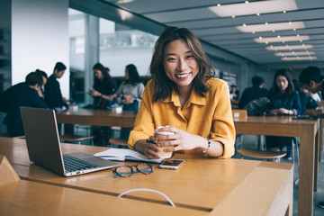 Portrait of happy female student sitting at desktop with laptop computer in university space feeling good during e learning, happy Japanese freelancer holding caffeine beverage and laughing indoors