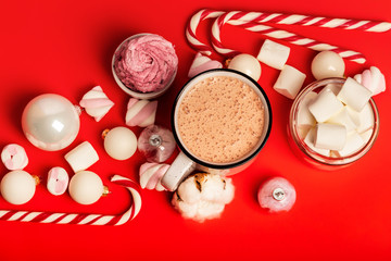 Cup of hot cocoa and marshmallows with christmas candy canes on a red background. White christmas tree decorations and candies.