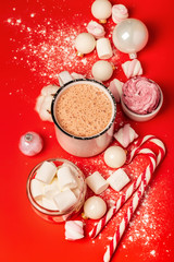 Cup of hot cocoa and marshmallows with christmas candy canes on a red background. White christmas tree decorations and candies.