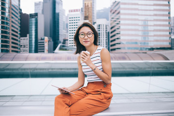 Fototapeta premium Portrait of Chinese female blogger with digital tablet for networking in hand looking at camera and smiling during free time at urban setting, positive woman in optical glasses for vision correction