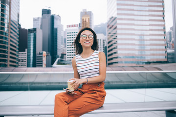 Fototapeta premium Portrait of Chinese female blogger with digital tablet looking at camera and smiling during free time at urban setting, positive woman in optical glasses for vision correction enjoying coffee time