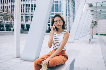 Portrait of attractive female student with takeaway cup sitting outdoors and calling to customer service for checking account bank balance, beautiful Japanese woman making international conversation