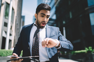 Turkish bearded male entrepreneur checking time on wearable mechanic wristwatch on way for business meeting with future partner shocked man in formal jacket feeling amazed from late to office