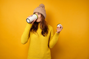 A brunette in a yellow sweater and hat stands on a yellow background and takes a sip from a white...