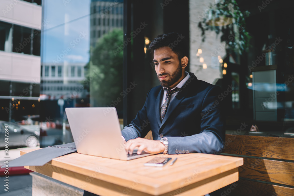 Poster spanish male economist working with report via application on laptop computer connected to cafeteria