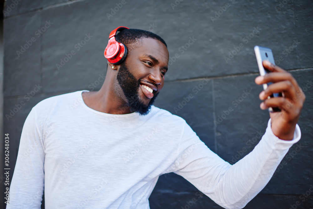 Aufkleber cheerful man smiling at camera while making selfie on smartphone standing on urban setting and enjoy
