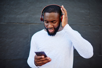 Dark skinned hipster guy updating music app on mobile phone to download audio via 4G internet connection, young man listening music playlist from cellular application via bluetooth headphones - obrazy, fototapety, plakaty