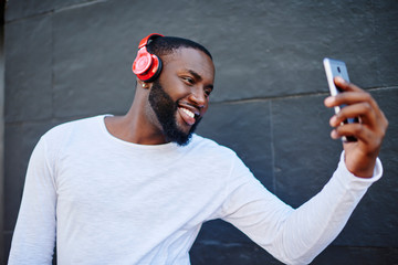 Cheerful man smiling at camera while making selfie on smartphone standing on urban setting and...
