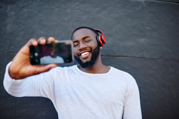 Happy millennial man blogger posing for smartphone camera shooting online video publication in earphones, smiling African American hipster guy taking selfie via cellular while enjoying music playlist