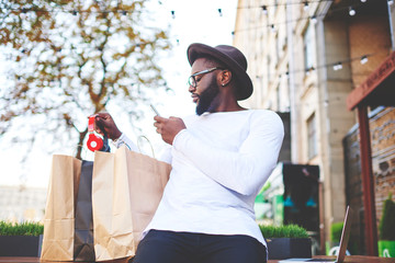 Dark skinned man checking purchases holding mobile phone and taking new bluetooth headphones for...