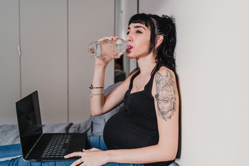 young pregnant woman at home working on laptop and drinking water