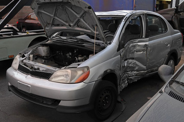 A generic passenger car with major damage to the driver side is shown in an automotive salvage yard during the late afternoon.