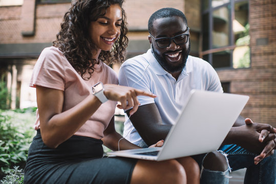 Happy Dark Skinned Colleagues Watching Comedy Movie Togetherness While Sitting Outdoors With Laptop Computer And Using One Electronic Headphones Both, Positive Couple In Love Laughing From Message