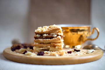 Piece of cake Napoleon on wooden plate, gold сup in distance
