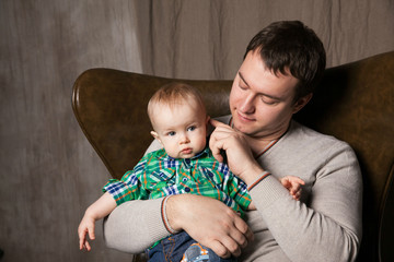 a young dad, 25 years old, of European appearance, sits in an armchair and gently holds his little son in his arms. Strong man’s shoulder, protection, happiness, calm