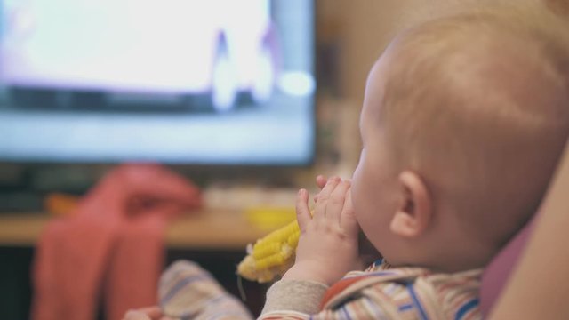 cute little child eats boiled corn on cob in mother arms watching tv in light room at home close side view