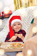 Fototapeta premium Little cute boy in santa hat ordered a big pizza from food delivery and eats it himself on the background of New Year's decorations.