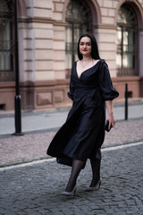 Fototapeta premium Autumn fashion outdoor. The girl with red lips in fashionable stylish black dress, autumnal lifestyle on the background of blurry yellow-green trees in the park. Vertical