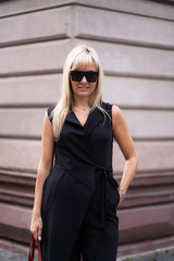 Autumn fashion outdoor. The blonde girl in fashionable stylish black overalls, sunglasses and red bag autumnal lifestyle on the background of blurry yellow-green trees in the park. Vertical