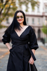 Autumn fashion outdoor. The girl with red lips in fashionable stylish black dress and sunglasses, autumnal lifestyle on the background of blurry yellow-green trees in the park. Vertical