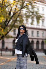 Fototapeta premium Autumn fashion outdoor. The girl in fashionable stylish white shirt , pants, jacket and sunglasses, autumnal lifestyle on the background of blurry yellow-green trees in the park. Vertical