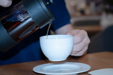 soft focus of man hands in blue t shirt sitting at table, pouring tea with glass kettle to white cup and smiling in cafe
