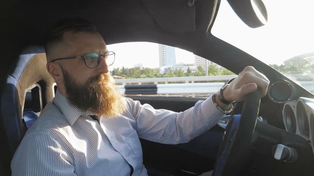 Serious young bearded man in eyeglasses sitting behind the wheel and driving sport car