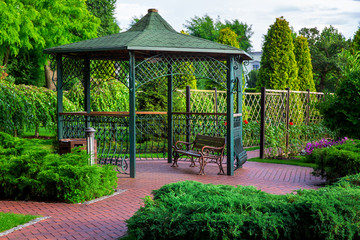 an iron gazebo with shingles and a park bench with bushes and trees, a lantern and an urn by the...