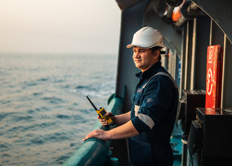 Marine Deck Officer or Chief mate on deck of offshore vessel or ship , wearing PPE personal protective equipment - helmet, coverall. He holds VHF walkie-talkie radio in hands.