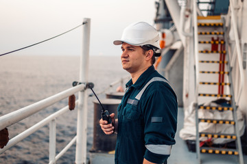 Marine Deck Officer or Chief mate on deck of offshore vessel or ship , wearing PPE personal protective equipment - helmet, coverall. He holds VHF walkie-talkie radio in hands.