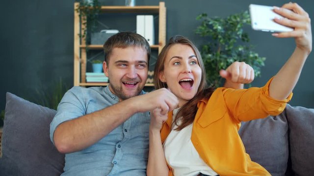 Happy young couple is taking selfie with smartphone at home gesturing posing sitting on couch togeher having fun. Relationship and technology concept.